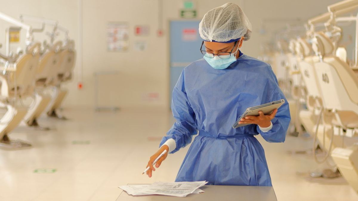 Petugas medis bermasker dan bergaun pelindung memeriksa formulir sambil memegang tablet